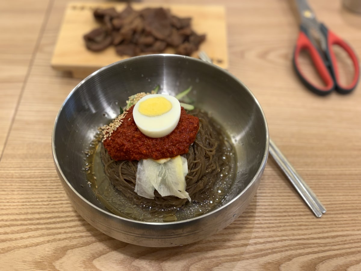 A bowl of Korean cold noodles with spicy red sauce, egg, cucumber, radish, broth, and grilled meat in the background.